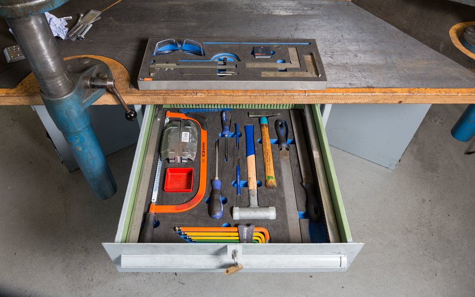 Das Bild zeigt eine geordnete Werkzeugbank. Mittig ist eine aufgezogene Schublade zu sehen, in der jedes Werkzeug seinen eigenen, sicheren Platz hat. Link zum Artikel.
