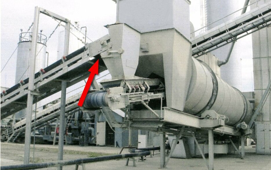 Das Foto zeigt eine Asphaltmischanlage mit Aufgabetrichter an einem Förderband in der Höhe. Ein roter Pfeil zeigt, von wo Material nach unten auf den Boden fiel und eine Reinigung erforderlich machte. Link zum Artikel.