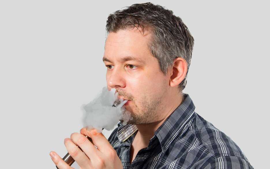 Das Foto zeigt einen Mann im Portrait, der E-Zigarette raucht und eine Dampfwolke aus Mund und Nase bläst. Link zur vergrößerten Darstellung des Bildes.