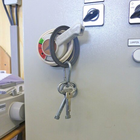 Das Bild zeigt einen Schlüsselbund, der mit einem Gummiring über den Hebel des Hauptschalters gehängt wurde. Hier kann sich jeder bedienen und die Anlage mit dem Schlüssel wieder einschalten. Link zur vergrößerten Darstellung des Bildes.