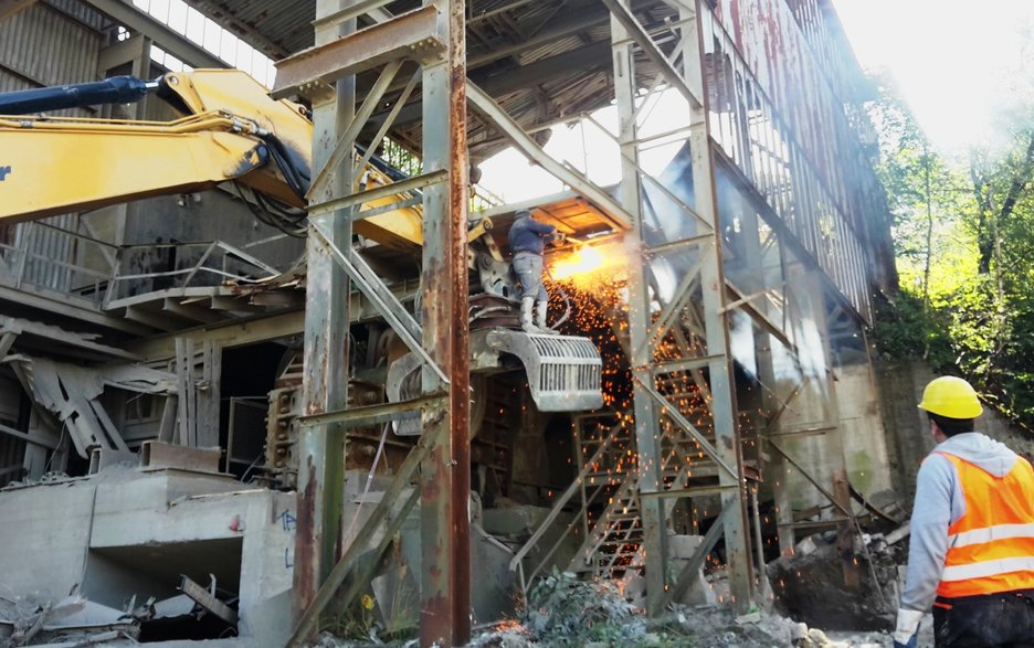 Das Bild zeigt ein Gebäude aus Stahl, das abgebrochen wird. Ein Mitarbeiter hat den Greifer eines Baggers in die Höhe gefahren und sich darauf gestellt, um in der Höhe zu schweißen. Eine halsbrecherische Aktion.  Link zum Artikel.