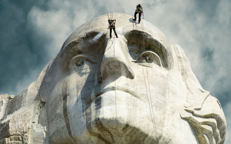 Das Foto zeigt das felsige Gesicht von George Washington von unten als Teil des US-amerikanischen Denkmals Mount Rushmore. Zwei Instandhalter lassen sich gerade an Sicherungsseilen an dem 18 Meter hohen Gesicht hinab, um es mit Hochdruckreinigern zu bearbeiten. Link zum Artikel.