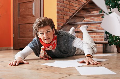 Das Bild zeigt eine Frau, die am Fuße einer Treppe auf dem Bauch liegt. Um sie herum liegen Papiere auf dem Boden verstreut. Es ist offensichtlich, dass sie die Treppe hintergestürzt ist. Link zur vergrößerten Darstellung des Bildes.