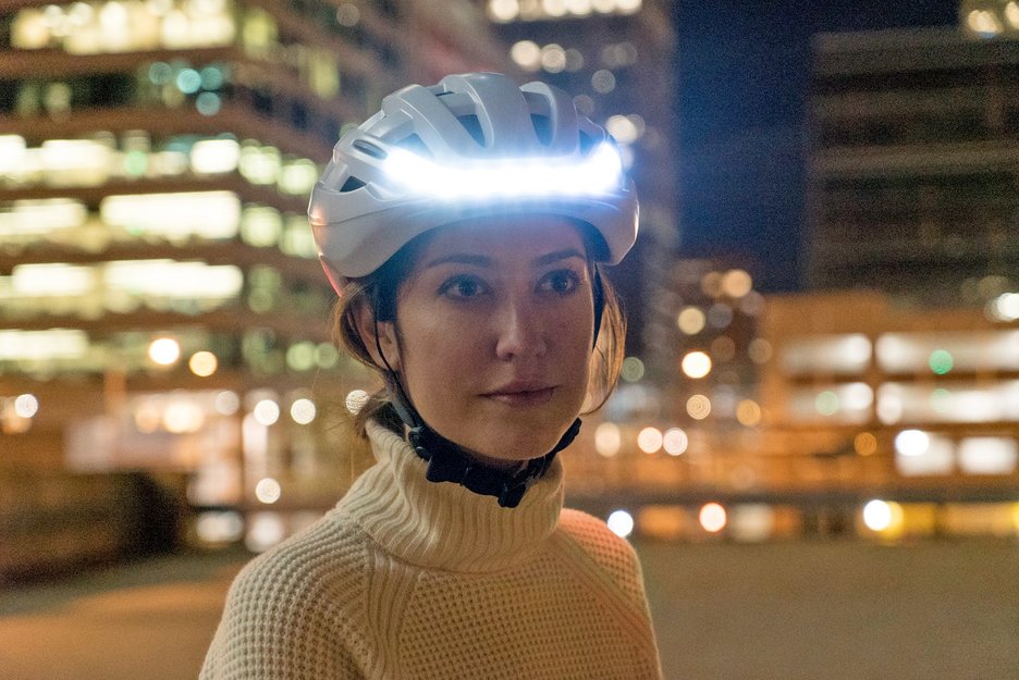 Das Bild zeigt eine Radfahrerin in einer Stadt bei Nacht. Sie trägt einen selbstleuchtenden Fahrradhelm und ist somit gut zu sehen. Link zur vergrößerten Darstellung des Bildes.
