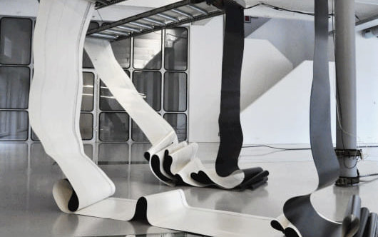 Zu sehen ist eine Bewegungsskulptur des Künstlers Zoro Feigl, die aus auf und ab tanzenden Gurten zweier Förderbänder besteht. Link zum Artikel.