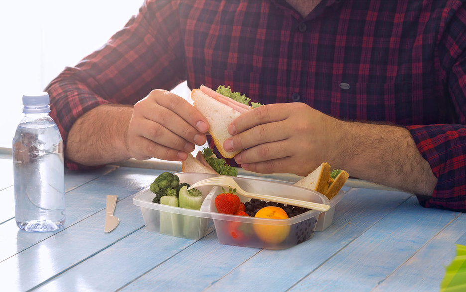Zu sehen ist der Oberkörper eines  Mannes, der am Tisch sitzt und isst. Vor ihm steht eine geöffnete Brotdose mit gesundem Obst und Gemüse. In den Händen hält er ein belegtes Sandwich. Auf der linken Seite des Bildes steht eine mit Wasser gefüllte Plastikflasche. Link zum Artikel.