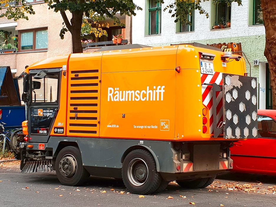 Das Bild zeigt eine moderne, orangefarbene Kehrmaschine der Berliner Stadtreinigung. Auf der Seite steht der witzige Name „Räumschiff“.  Link zur vergrößerten Darstellung des Bildes.