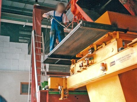 Zu sehen ist ein Mitarbeiter in großer Höhe auf einer ungesicherten Plattform an einer Kübelbahn. Er arbeitet mit einem Hochdruckreiniger. Hier besteht akute Absturzgefahr. Link zur vergrößerten Darstellung des Bildes.