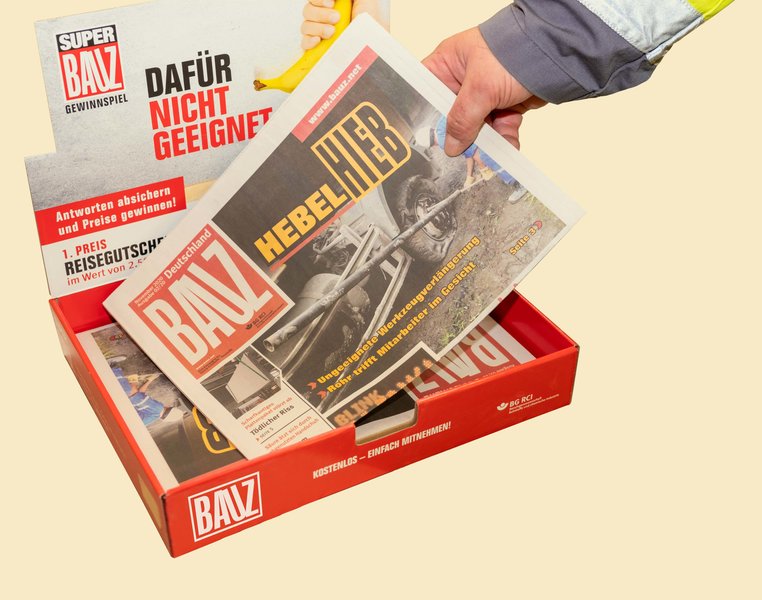 Das Bild zeigt einen BAUZ-Tresenaufsteller und eine Hand, die eine der darin liegenden BAUZ-Zeitungen nimmt.