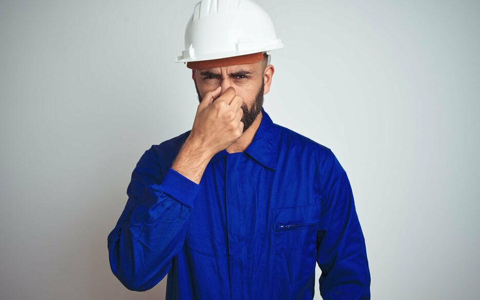 Das Symbolbild zeigt einen Mitarbeiter frontal im Portrait mit weißem Helm und blauem Hemd. Er hält sich die Nase zu, so als ob etwas unangenehm riechen würde. Das Bild unterstützt die grundsätzliche Frage, ob die Nase tatsächlich vor Gefahrstoffen warnen kann. Link zum Artikel.