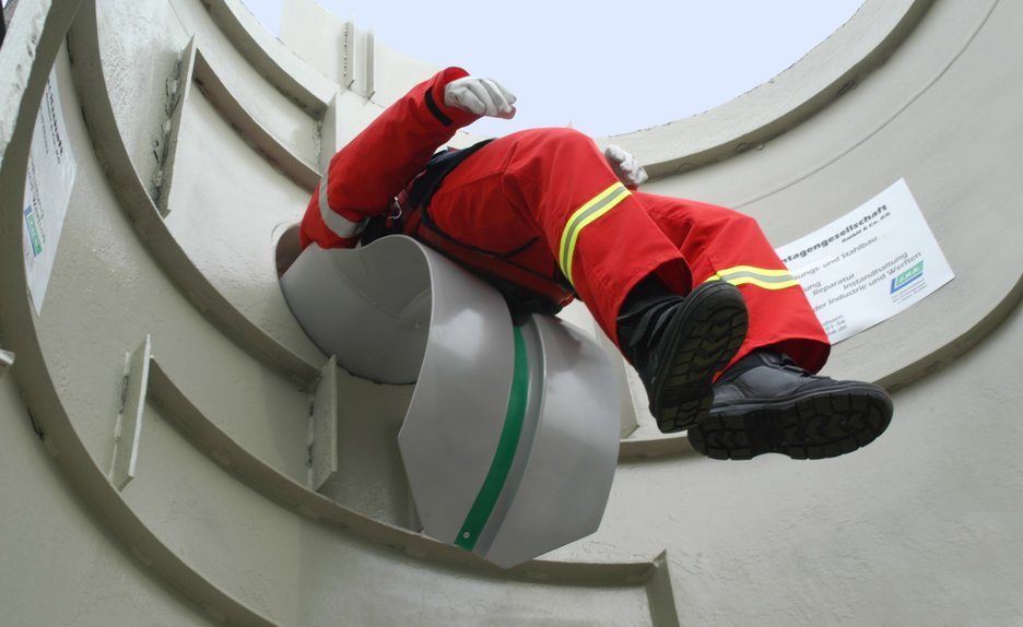 Das Bild zeigt, wie ein Mitarbeiter mithilfe einer Rettungsrutsche aus einem Silo geborgen wird. Die Rettungsrutsche sieht aus wie ein großer Schuhlöffel, auf dem der Verletzte aus dem Silo gleitet. Link zur vergrößerten Darstellung des Bildes.