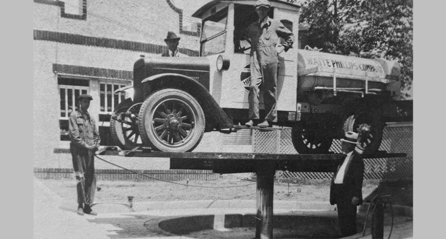 Das Schwarz-Weiß-Foto zeigt eine Erfindung des US-amerikanischen Automechanikers Peter Lunati: die hydraulische Hebebühne. Zu sehen ist, wie diese einen alten Tankwagen anhebt. Der Erfinder steht rechts daneben und blickt auf den Wagen. Ein Mann steht auf dem Einstieg des Wagens, ein anderer seitlich neben der Bühne und hält ein Seil. Ein dritter Mann mit weißem Hut schaut hinter dem Wagen hervor. Link zur vergrößerten Darstellung des Bildes.