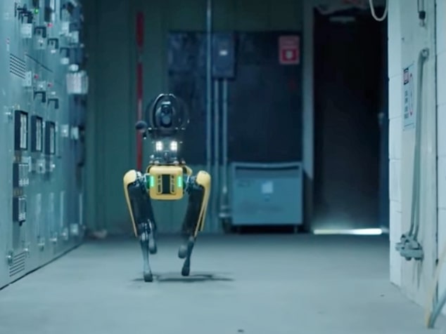 Das Bild zeigt den Roboter-Hund Spot von vorne in einer dunklen Werkhalle zwischen Schaltschränken. Er navigiert autonom als Nachtwächter durch eine Fabrik. Bei seinem Kontrollgang stellt er fest, ob Türen geschlossen sind oder ob sich noch Personen in einem Raum aufhalten. Link zur vergrößerten Darstellung des Bildes.
