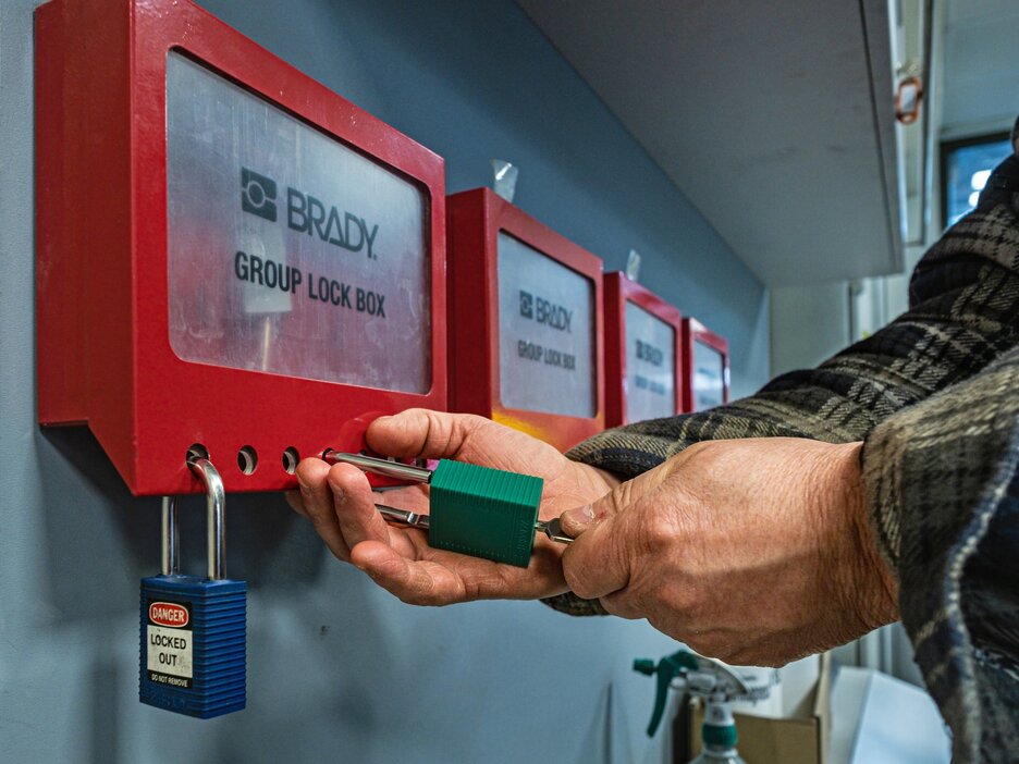 Sicherungsbox für Anlagenschlüssel Das Bild zeigt eine Reihe roter Sicherungsboxen, die an einer Wand hängen. Am unteren Rand sind Löcher zu sehen, in denen Schlösser eingehängt werden können. Ganz links hängt bereits ein schwarzes Schloss. Rechts sind die Hände eines Mitarbeiters zu sehen, der gerade ein Schloss in eines der Löcher eingehängt hat und dieses abschließt. In der roten Box liegt ein Schlüssel. Dieser gehört zu dem Schloss, mit dem eine stillgesetzte Anlage gesichert wird. Den Schlüssel gibt es nur einmal. Bei Reparaturen wird dieser einzige Schlüssel aus Sicherheitsgründen vom Schloss abgezogen und in der Box hinterlegt. Jeder Mitarbeitende, der an der Anlage tätig ist, muss hier sein persönliches Sicherheitsschloss einhängen und abschließen. Die Box kann somit nicht versehentlich geöffnet werden. Der Anlagenschlüssel befindet sich gesichert so lange in der Box, bis der letzte Mitarbeitende sein Schloss wieder entfernt hat. Link zur vergrößerten Darstellung des Bildes.