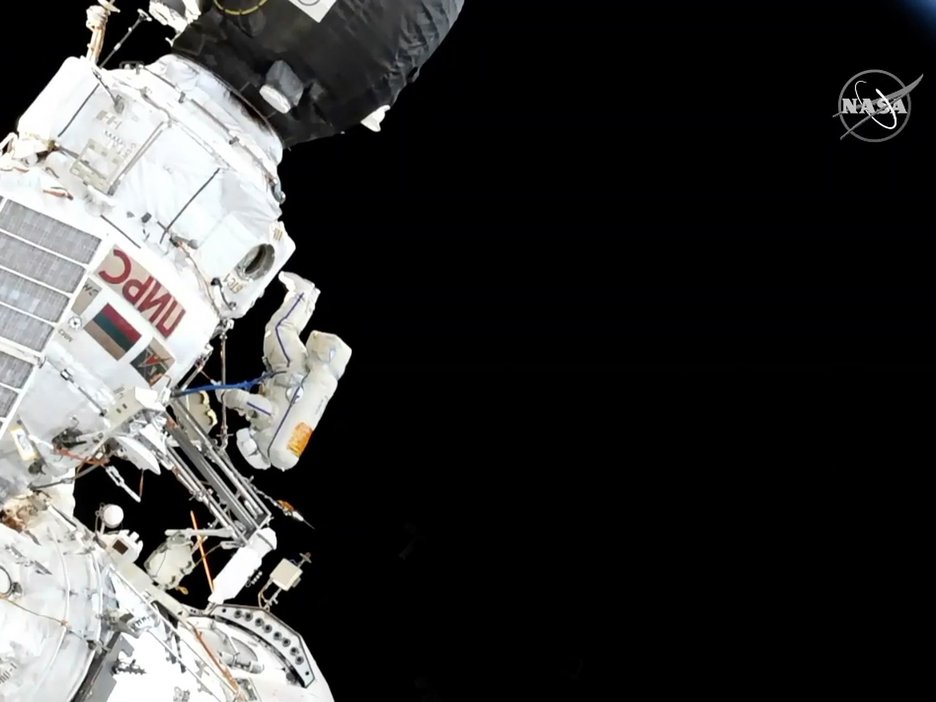 Das Bild zeigt die Raumstation ISS von außen im Weltall und einen Astronauten, der kopfüber schwebt und von außen Reinigungsarbeiten durchführt.  