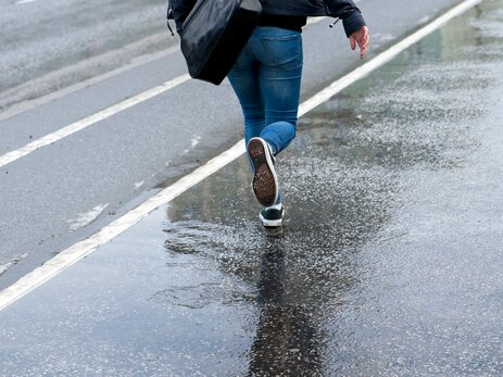Das Foto zeigt den Ausschnitt einer Fußgängerin von hinten, die über eine nasse Straße läuft. Link zur vergrößerten Darstellung des Bildes.