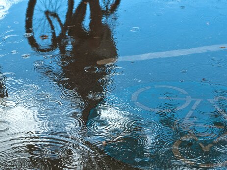 Das Foto zeigt die Spiegelung eines Radfahrers in einer Pfütze auf der Straße, während es regnet. Hier geht es um schlechtes Sehen und Gesehenwerden. Dies ist ein Risiko bei Unwetter für Zweiradfahrer im Straßenverkehr. Link zur vergrößerten Darstellung des Bildes.