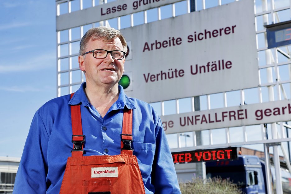 Zu sehen ist der Leiter der mechanischen Werksstatt Jürgen Riedemann vor einem Schild mit der Aufschrift „Arbeite sicherer, verhüte Unfälle“. Link zur vergrößerten Darstellung des Bildes.