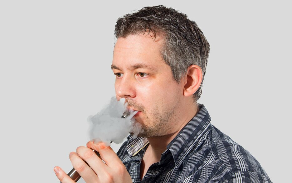 Das Foto zeigt einen Mann im Portrait, der E-Zigarette raucht und eine Dampfwolke aus Mund und Nase bläst.