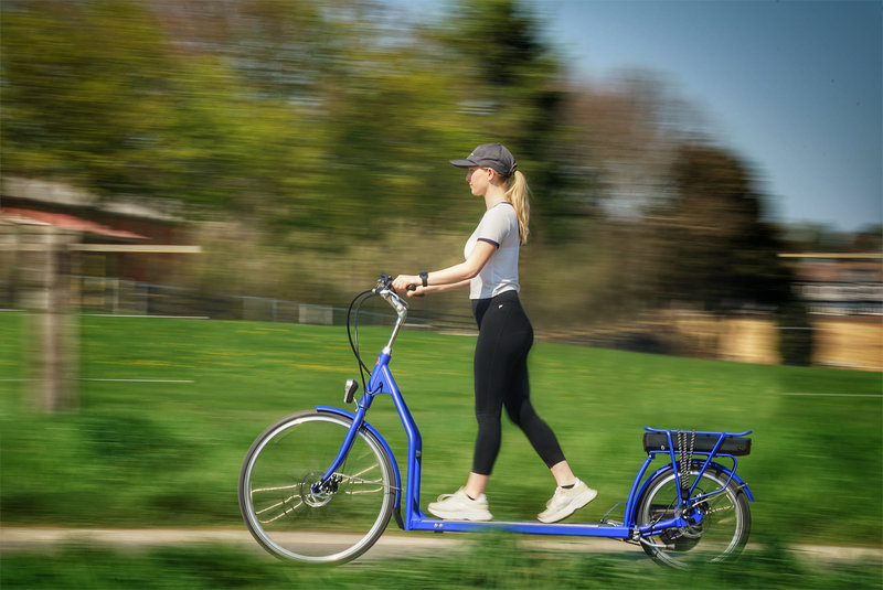 Zu sehen ist eine junge Frau auf einem langen blauen Fahrrad. Sie läuft in der Mitte des Rades mit den Füßen vor und zurück, denn hier befindet sich ein Laufband, welches das Fahrrad antreibt.