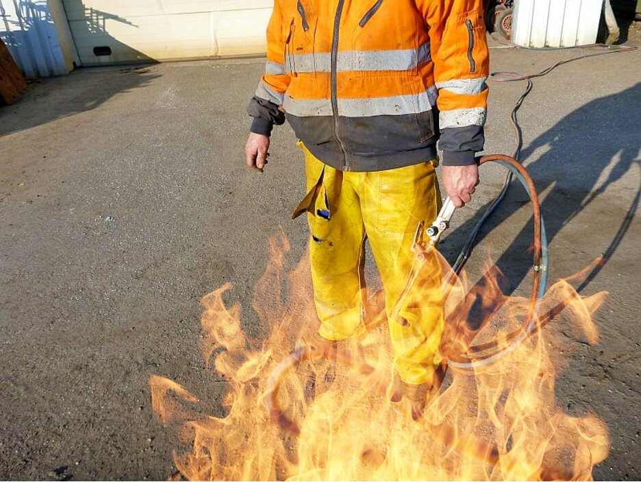 Das Bild zeigt eine Bildmontage. Zu sehen sind Bauch und Beine eines Mitarbeiters, der einen Schweißbrenner in der linken Hand hält. Oben trägt er eine Wetterschutzjacke und unten eine gelbe Ölhose. Grafische Flammen an den Beinen sollen den nachgestellten Unfall verdeutlichen. Link zur vergrößerten Darstellung des Bildes.