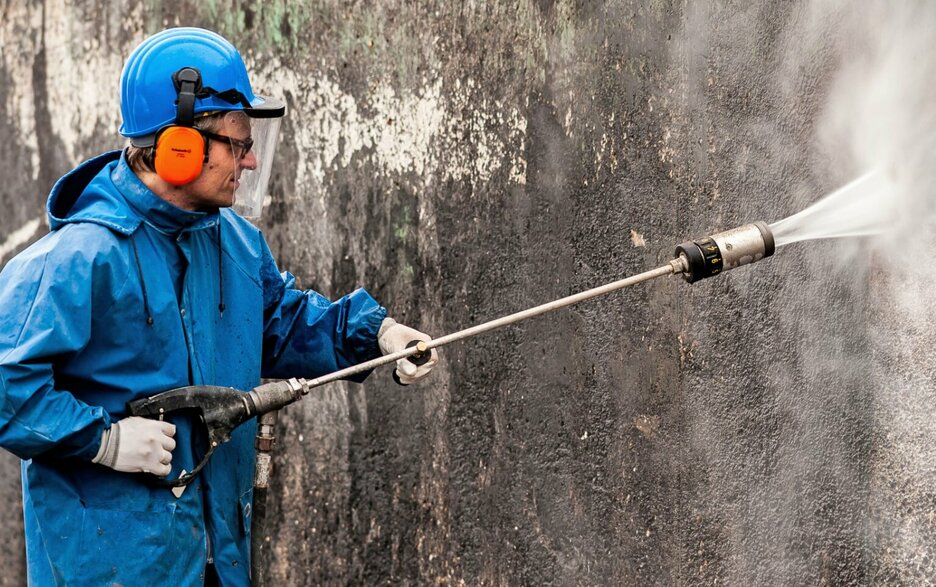 Zu sehen ist ein Mitarbeiter in geeigneter Persönlicher Schutzausrüstung, der mit einem Hochdruckreiniger eine Wand mit dem harten Wasserstrahl säubert. 