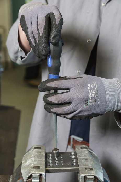 Das Bild zeigt zwei Hände, die Schutzhandschuhe tragen und mit einem Schraubendreher an einem eingespannten Werkstück arbeiten. Beide Hände führen den Schraubendreher. Link zur vergrößerten Darstellung des Bildes.