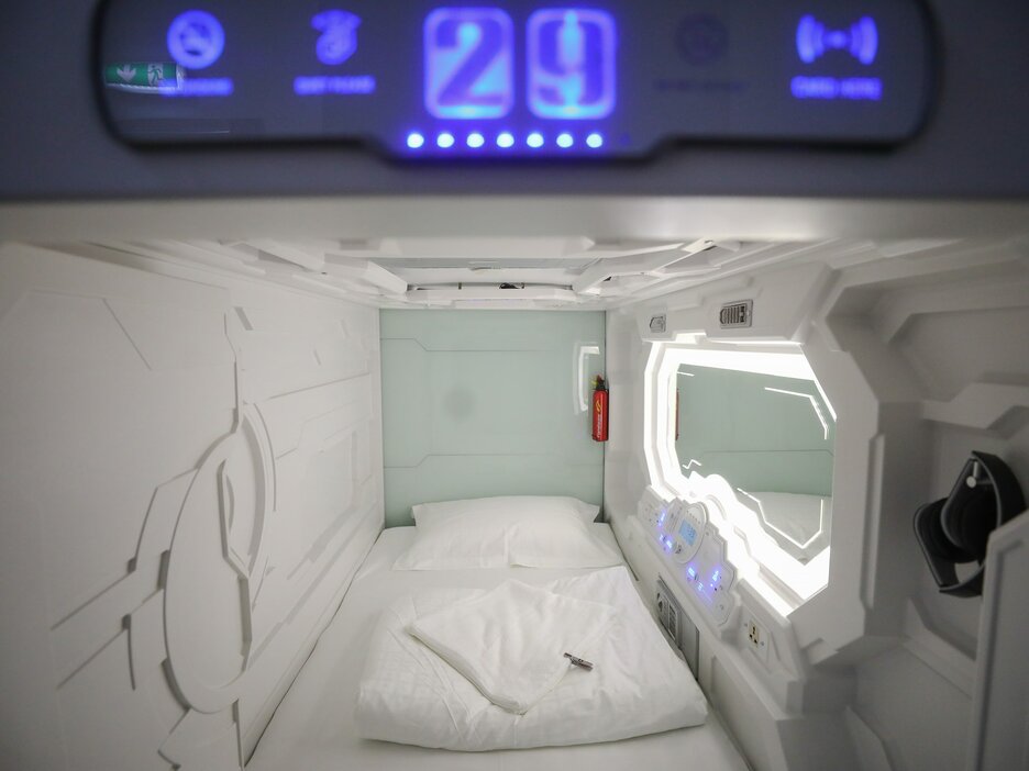 Das Bild zeigt den Blick in eine futuristisch wirkende fensterlose Schlafkapsel. Dabei handelt es sich um ein preiswertes, fensterloses Mini-Hotelzimmer für Reisende mit geringerem Budget und ohne Platzangst. Es besteht lediglich aus einem Bett und einem Spiegel an der rechten Wand. Unter dem Spiegel sind eine Uhr und Bedienelemente mit LEDs zu sehen. Link zur vergrößerten Darstellung des Bildes.