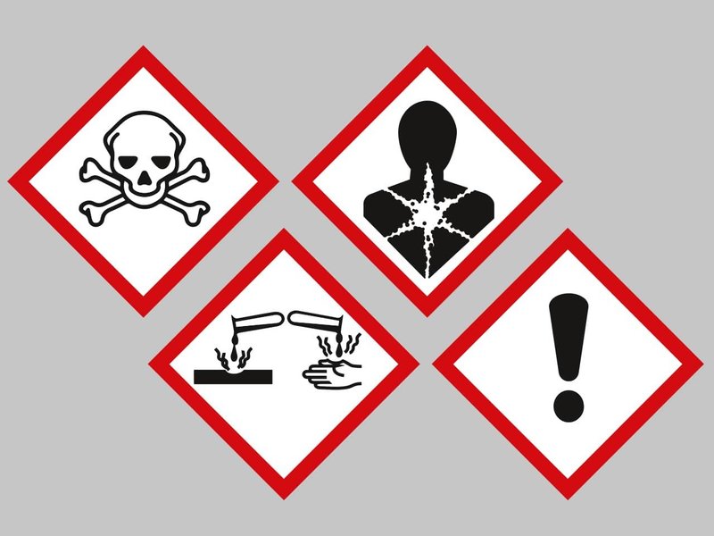 Zu sehen sind vier verschiedene Gefahrstoffzeichen. Ein Totenkopf für giftig, eine Person mit sternförmig aufgerissenem Oberkörper für Gesundheitsgefahr, zwei tropfende Reagenzgläser mit einer Hand für ätzend, ein Ausrufezeichen für reizend. 