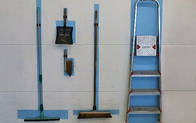 Das Bild zeigt eine weiße Wand, auf der blaue Markierungen zu sehen sind. Die Markierungen haben die Größe und Form des jeweiligen Arbeitsmittels, das dorthin zurückgehängt werden soll. Hier befinden sich gerade ein Schrubber, ein Kehrblech, ein Handfeger, ein Besen und eine Leiter an ihrem richtigen Platz an der Wand. Zur so genannten 5S-Methode, bei der Arbeitsplätze aufgeräumt, unnötiges aussortiert, alles sauber gehalten und markiert wird, gehören feste Plätze für Arbeitsmittel. In diesem Fall geht es um Leitern. Link zum Artikel.