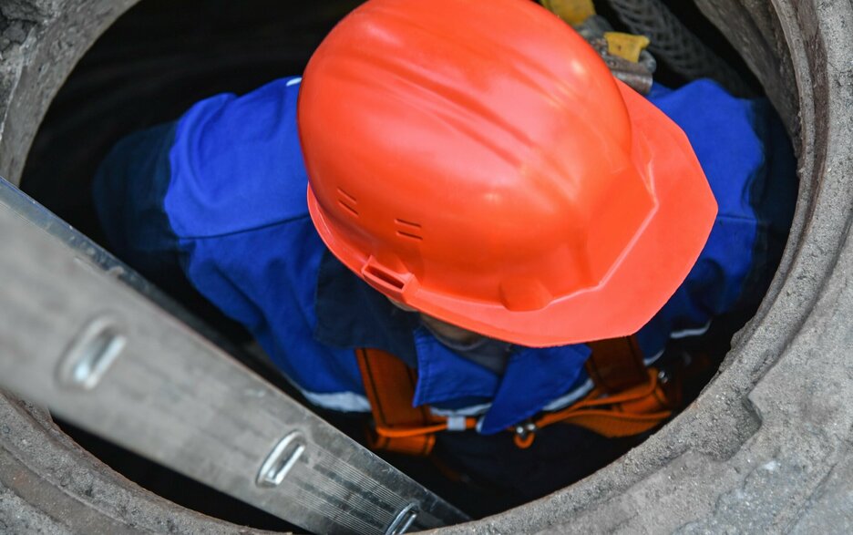 Das Bild zeigt den Blick auf einen Mitarbeiter mit rotem Helm von oben, der gesichert über eine Leiter in einen engen Schacht hinabsteigt. Es handelt sich hierbei um ein Symbolbild, was das Thema „enger Raum“ verdeutlichen soll.  Link zum Artikel.