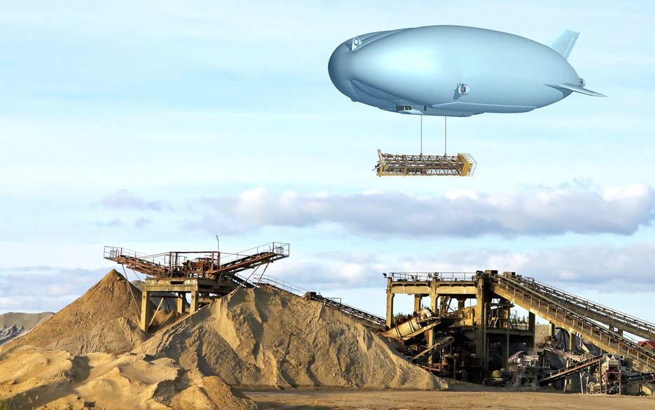 Das Bild zeigt einen Zeppelin, unter dem eine große Last hängt. Dieser schwebt über einer Baustelle in unwegsamem Gelände, an der gerade eine Brücke entsteht. Der Lasten-Zeppelin kann Teile mit bis zu neunzig Tonnen Gewicht heben.