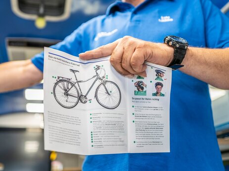 Das Foto zeigt einen Mitarbeiter im VISION ZERO-Ideenmobil, der einen Flyer mit einem Fahrrad hochhält. Anhand von Infomaterial wird das Thema „Verkehrssicheres Fahrrad“ vertieft. Link zur vergrößerten Darstellung des Bildes.