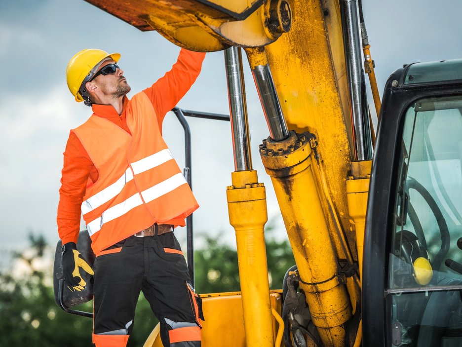 Auf dem Foto ist ein Mitarbeiter neben einer Erdbaumaschine zu sehen. Er prüft gerade etwas an der Hydraulik an dem Baggerarm über ihm und trägt dabei einen gelben Helm, Sonnenbrille mit UV-Schutz, eine orangene Warnweste und schwarz-gelbe Schutzhandschuhe.