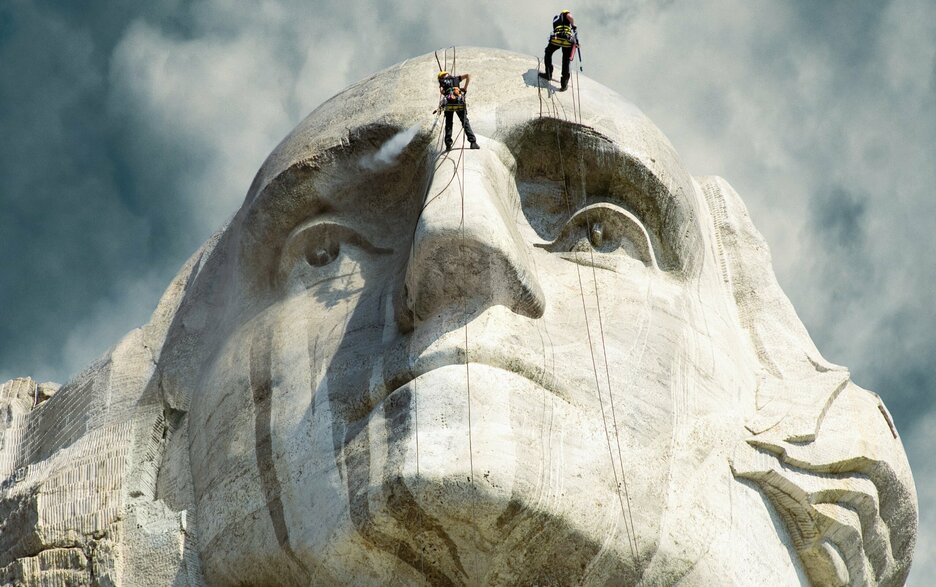 Das Foto zeigt das felsige Gesicht von George Washington von unten als Teil des US-amerikanischen Denkmals Mount Rushmore. Zwei Instandhalter lassen sich gerade an Sicherungsseilen an dem 18 Meter hohen Gesicht hinab, um es mit Hochdruckreinigern zu bearbeiten.