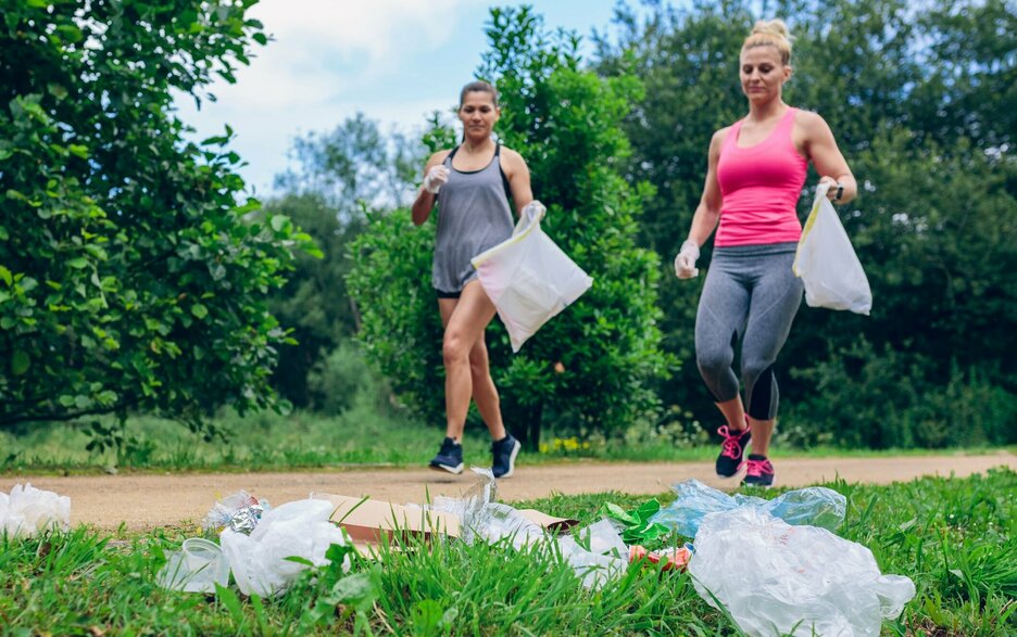 Das Bild zeigt im Bildhintergrund zwei Frauen in Sportoutfit, die beim Joggen in der Natur Handschuhe und jeweils eine Mülltüte tragen. Im Bildvordergrund liegt Müll auf einer grünen Wiese. Beide Frauen laufen darauf zu, um diesen einzusammeln. Das Bild verdeutlicht eine sportliche Aktivität, die sich Plogging nennt. Dies bedeutet zu joggen und gleichzeitig Müll aufzusammeln für eine saubere Umwelt. Link zum Artikel.