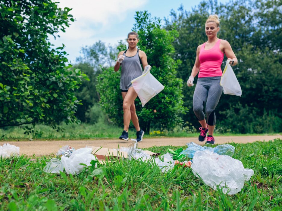 Das Bild zeigt im Bildhintergrund zwei Frauen in Sportoutfit, die beim Joggen in der Natur Handschuhe und jeweils eine Mülltüte tragen. Im Bildvordergrund liegt Müll auf einer grünen Wiese. Beide Frauen laufen darauf zu, um diesen einzusammeln. Das Bild verdeutlicht eine sportliche Aktivität, die sich Plogging nennt. Dies bedeutet zu joggen und gleichzeitig Müll aufzusammeln für eine saubere Umwelt. Link zur vergrößerten Darstellung des Bildes.