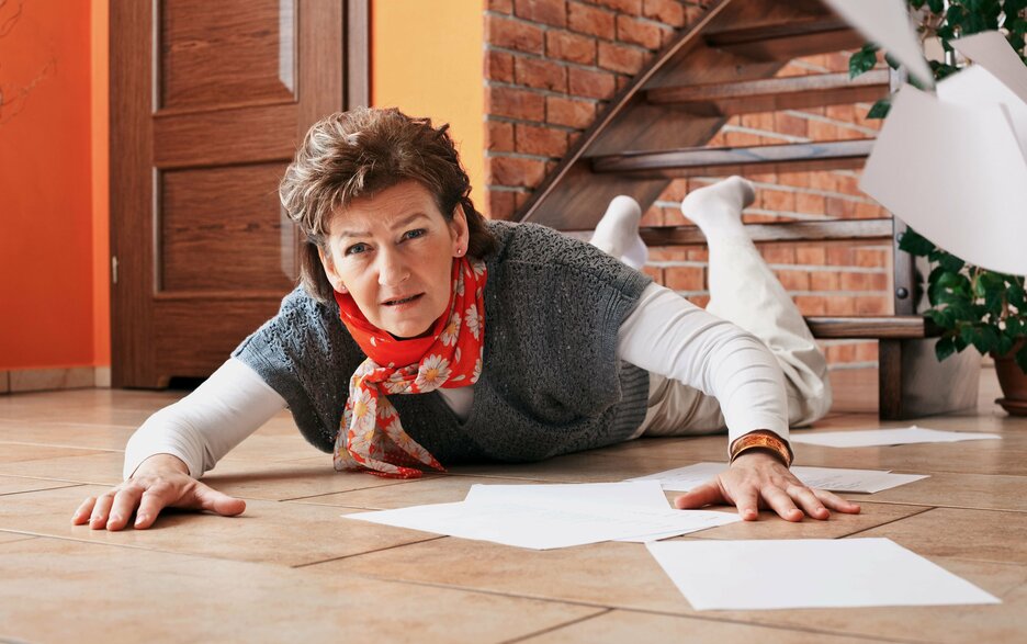 Das Bild zeigt eine Frau, die am Fuße einer Treppe auf dem Bauch liegt. Um sie herum liegen Papiere auf dem Boden verstreut. Es ist offensichtlich, dass sie die Treppe hintergestürzt ist. Link zum Artikel.