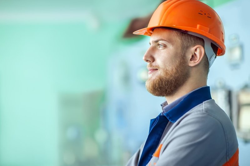 Zu sehen ist das Symbolbild eines nachdenklichen Mitarbeiters in Arbeitskleidung und mit orangefarbenem Helm. Er steht für das Thema „Arbeiten mit Diabetes“.
