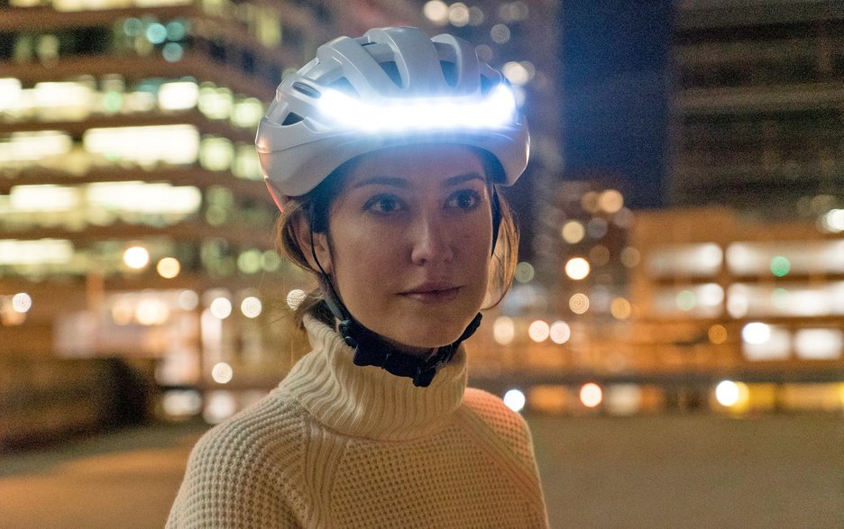 Das Bild zeigt eine Radfahrerin in einer Stadt bei Nacht. Sie trägt einen selbstleuchtenden Fahrradhelm und ist somit gut zu sehen.