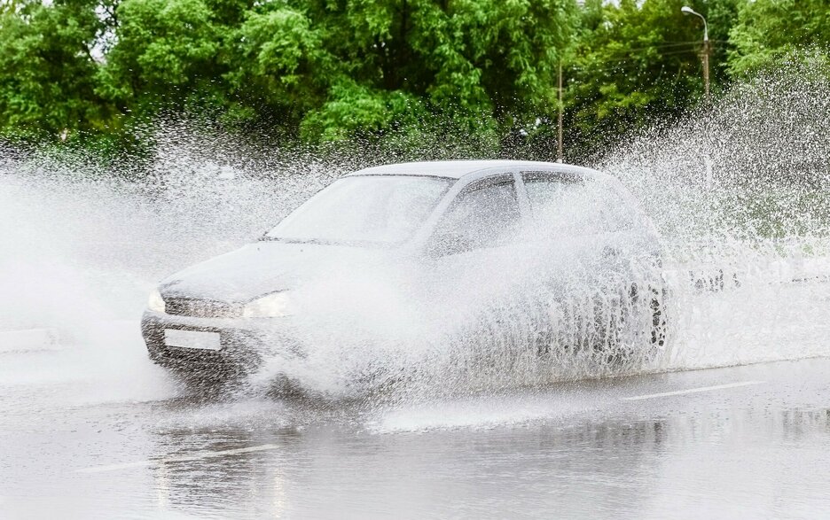 Das Foto zeigt ein Auto, das bei einem Starkregenguss durch stehendes Wasser auf der Fahrbahn fährt. Links und rechts vom Auto spritzt das Wasser meterweit hoch. Hier besteht die Gefahr von Aquaplaning. Link zum Artikel.