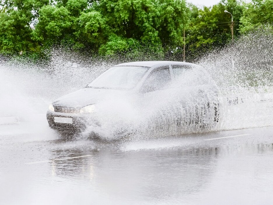 Das Foto zeigt ein Auto, das bei einem Starkregenguss durch stehendes Wasser auf der Fahrbahn fährt. Links und rechts vom Auto spritzt das Wasser meterweit hoch. Hier besteht die Gefahr von Aquaplaning. Link zur vergrößerten Darstellung des Bildes.