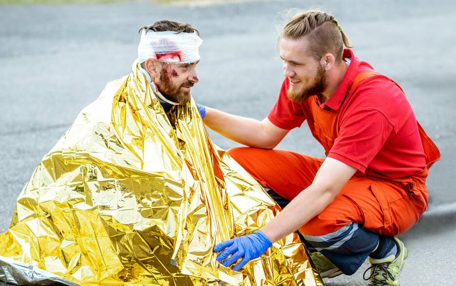 Symbolfoto: Verletzter Mitarbeiter nach Brand in Rettungsdecke Das nachgestellte Bild zeigt eine Unfallsituation. Zu sehen ist ein verletzter Mann, der in eine goldfarbige Rettungsdecke gehüllt auf dem Boden sitzt. Am Kopf trägt er einen blutigen Verband. An seiner rechten Wange ist eine Wunde zu sehen. Ein Rettungssanitäter kniet neben ihm und kümmert sich um ihn.