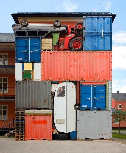 Das Bild zeigt eine riesige Skulptur des schwedischen Künstlers Michael Johansson. Dieser stapelt große, schwere Objekte und setzt sie zu einer Gesamtskulptur neu zusammen. Zu sehen sind hier ein Auto über Kopf, ein Traktor und ein Wohnwagen hochkant, eine grüne Mülltonne und mehrere farbige Container als rechteckige Einheit.