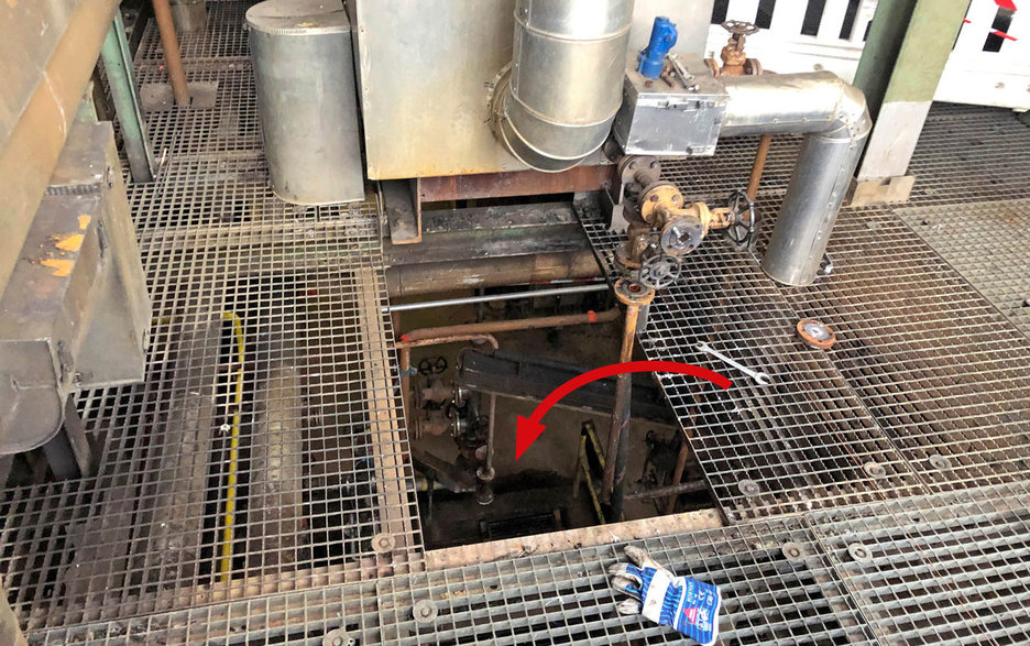 Das Bild zeigt die Absturzstelle in der Höhe an einem Wärmetauscher. Ein roter Pfeil weist auf ein Loch in den Gitterrostbelägen hin. Hier stürzte ein Mitarbeiter hindurch, weil ein Gitterrost nur lose aufgelegt war.