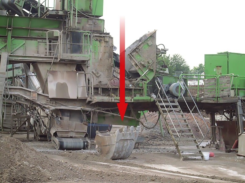 Das Bild zeigt eine Prallmühle, an der gearbeitet wurde und eine abgestürzte Baggerschaufel, die nicht richtig verriegelt war. Ein roter Pfeil weist daraufhin, wo die Schaufel hinunterkrachte.