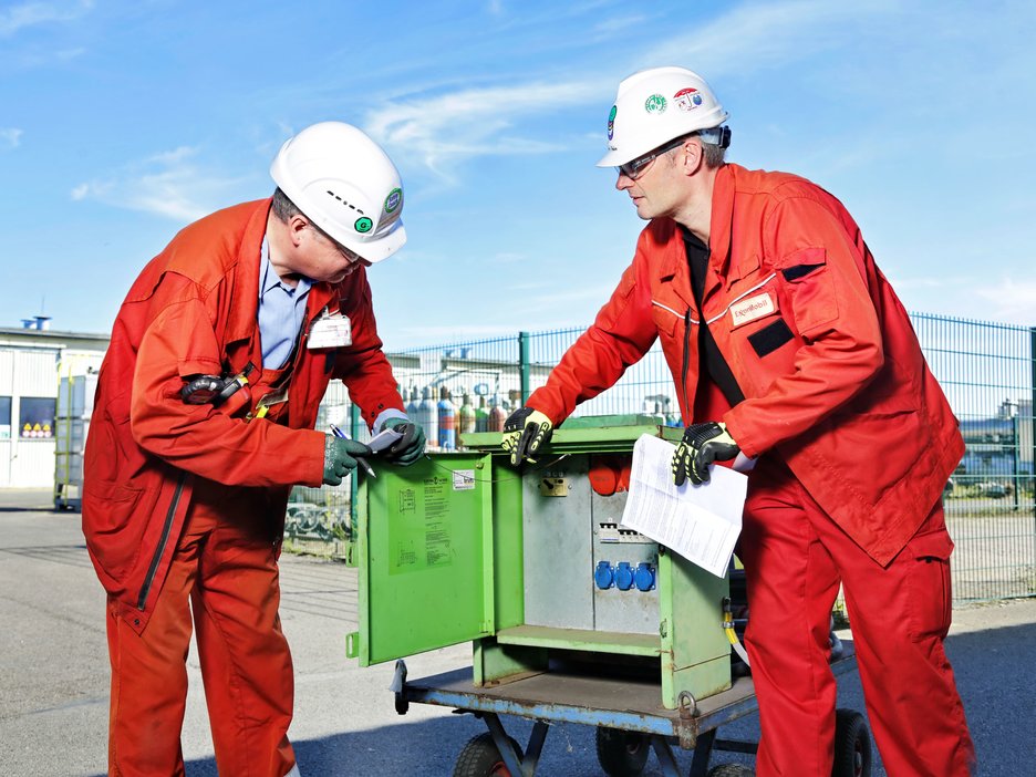 Das Bild zeigt den Safety-Coach Joachim Reimann und seinen Kollegen Christian Hahn, die sich über einen offenen Stromkasten beugen und gemeinsam eine Gefährdungsbeurteilung erstellen.  Link zur vergrößerten Darstellung des Bildes.