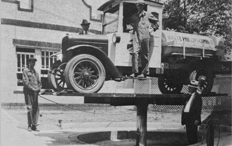Das Schwarz-Weiß-Foto zeigt eine Erfindung des US-amerikanischen Automechanikers Peter Lunati: die hydraulische Hebebühne. Zu sehen ist, wie diese einen alten Tankwagen anhebt. Der Erfinder steht rechts daneben und blickt auf den Wagen. Ein Mann steht auf dem Einstieg des Wagens, ein anderer seitlich neben der Bühne und hält ein Seil. Ein dritter Mann mit weißem Hut schaut hinter dem Wagen hervor. Link zum Artikel.