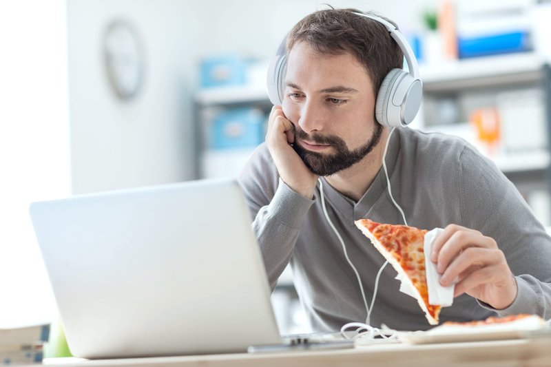 Zu sehen ist ein Mann mit Kopfhörern vor seinem Laptop. Gebannt schaut er auf den Bildschirm, während er seinen Kopf auf seinen linken Arm stützt. Mit der rechten Hand hält er ein Stück Pizza, das noch unberührt ist. Dieses Symbolfoto soll die Abhängigkeit und das sich Verlieren in der digitalen Welt von Online-Süchtigen verdeutlichen.