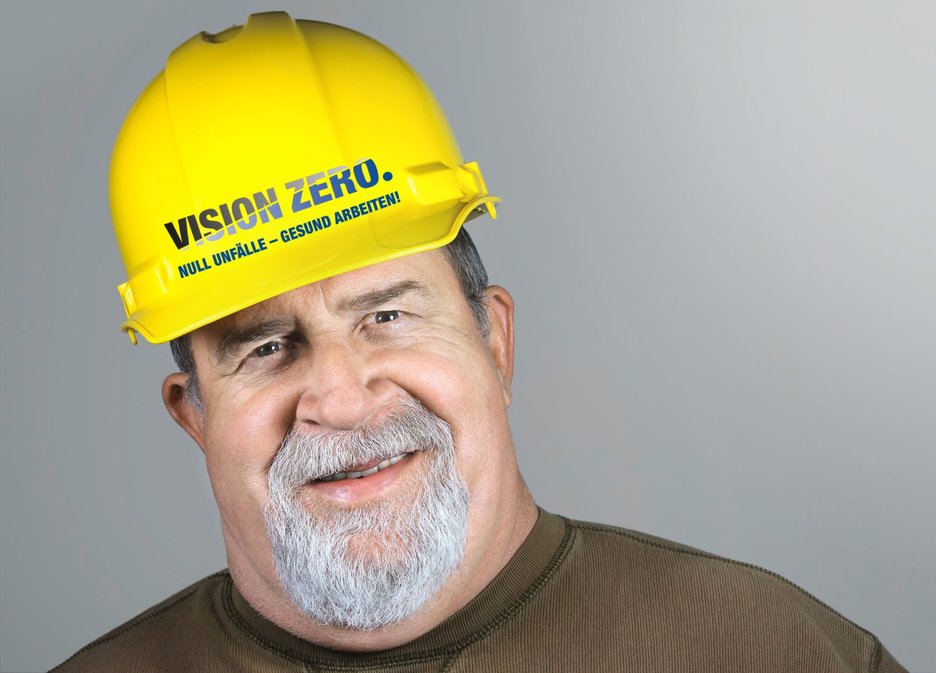 Das Bild zeigt einen lächelnden Bauarbeiter. Auf seinem Helm ist VISION ZERO zu lesen ist. Dieses Zeichen steht für null Unfälle im Betrieb. Link zur vergrößerten Darstellung des Bildes.
